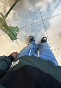 Person iført grønn og svart hettegenser, blå jeans og svarte sko som står på flislagt gulv nær et skadet grønt planteløv.