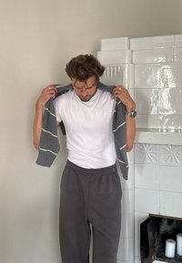 Cárdigan gris rayado sostenido sobre una camiseta blanca, combinado con pantalones de chándal grises. El cabello está despeinado y lleva un reloj en la muñeca izquierda.