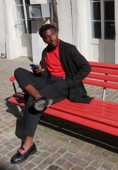 Suéter de punto rojo, cárdigan negro, pantalones negros y mocasines negros con acentos rojos. Sentado en un banco rojo contra un fondo de pavimento de piedra.