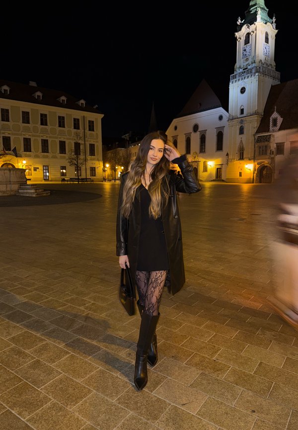 Mladá žena s dlouhými vlasy v černých šatech, koženém kabátě, vzorovaných punčochách a botách stojí na osvětleném náměstí ve městě v noci poblíž historických budov.
