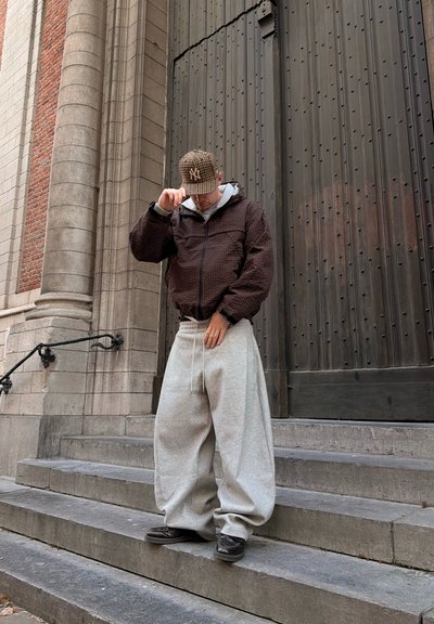 Hombre con pantalones deportivos grises extragrandes y chaqueta marrón ajustándose una gorra a cuadros mientras se encuentra en escaleras de piedra junto a una gran puerta de madera.