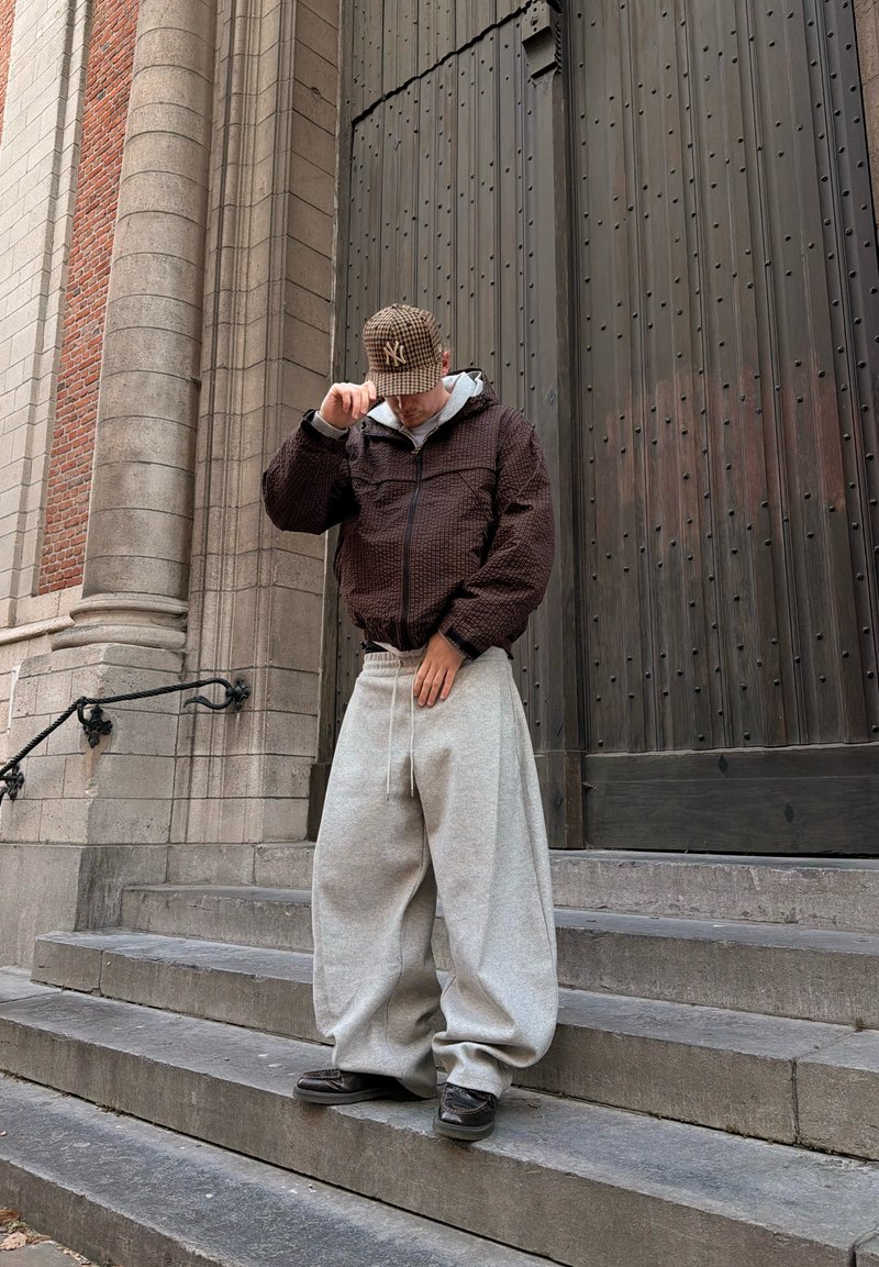 Homme en pantalon de survêtement gris surdimensionné et veste marron ajustant une casquette à carreaux, debout sur des marches en pierre près d'une grande porte en bois.