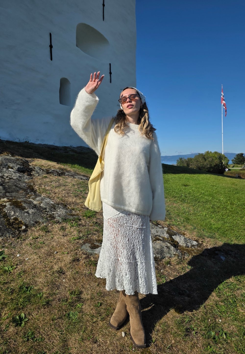 Suéter blanco y esponjoso, falda blanca de encaje, botas altas de color tostado. Los accesorios incluyen gafas de sol y un bolso amarillo. Entorno al aire libre con piedras y hierba.