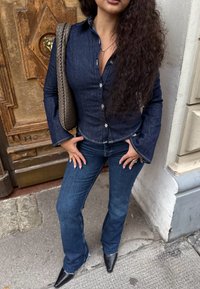 Dunkelblaues Hemd mit langen Ärmeln, kombiniert mit dunklen, ausgestellten Jeans. Accessoires sind eine geflochtene graue Tasche und spitze schwarze High Heels.