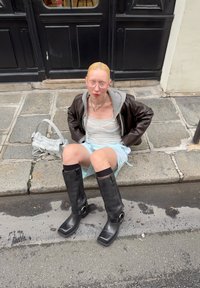 Schwarze lederne kniehohe Stiefel mit quadratischen Zehen und Schnallen-Details, kombiniert mit einem hellen Tanktop und Jeansshorts, sitzend auf einer Kopfsteinpflasterstraße.