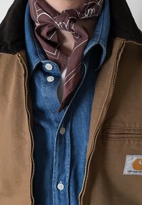 Καφέ μπουφάν Carhartt πάνω από μπλε τζιν πουκάμισο με λευκά κουμπιά, συνδυασμένο με μπουργουνδί φουλάρι με σχέδιο δεμένο γύρω από τον λαιμό.