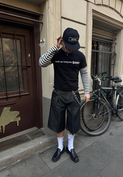 Camiseta negra con texto blanco, mangas largas a rayas, pantalones cortos de mezclilla oscura con detalle de cadena, calcetines blancos y mocasines negros.