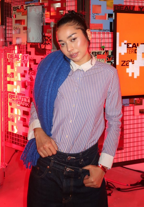 Kvinde med blå tørklæde over skulderen, iført stribet skjorte med knapper og jeans, poserer foran et skilt til et skønhedsevent oplyst med rødt lys.