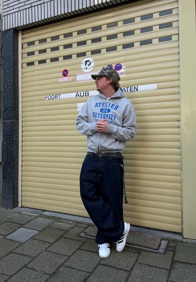 Hombre con sudadera gris y gorra apoyado contra una puerta de garaje beige cerrada con señales de advertencia, de pie sobre una acera con baldosas.