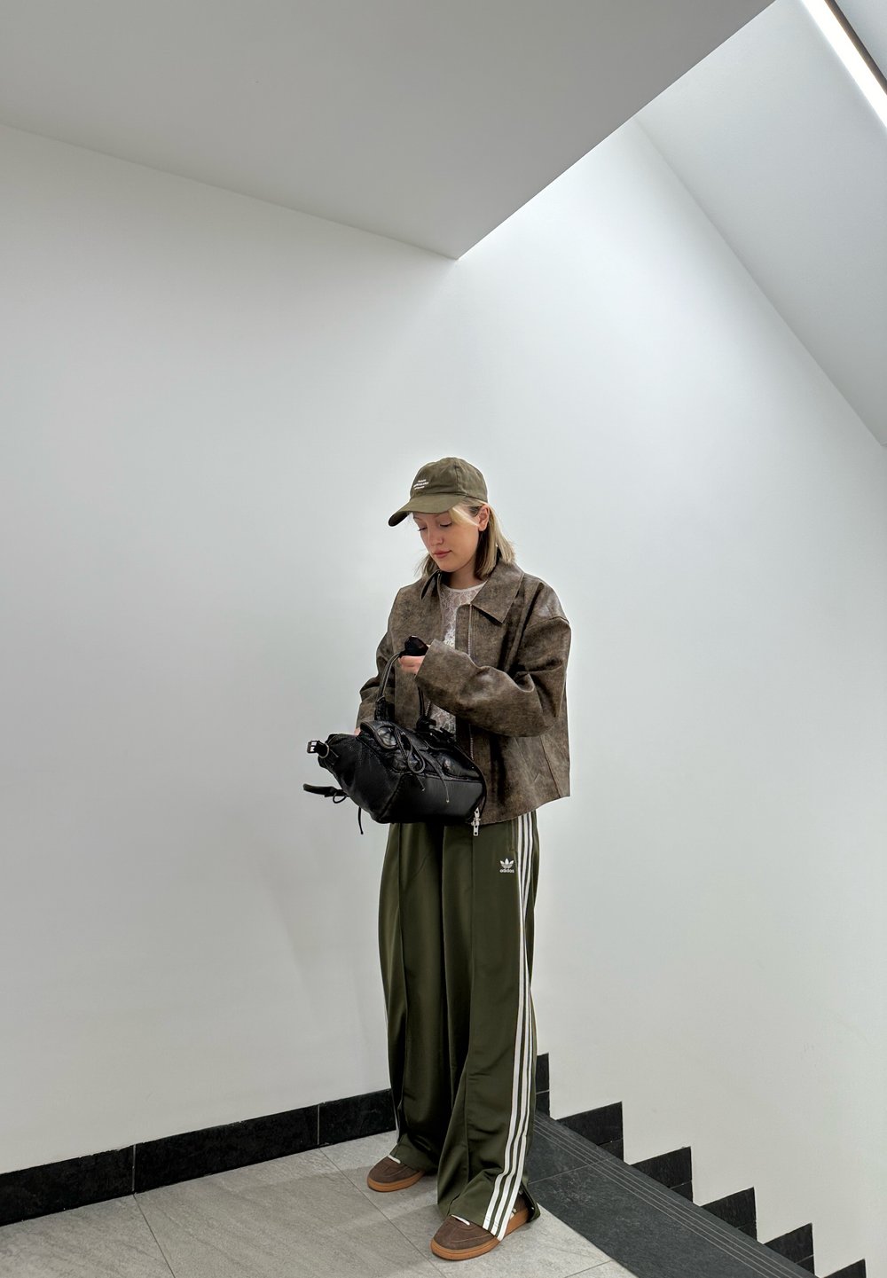 Καφέ κοντό σακάκι, ελαιόχρωμο παντελόνι με άσπρες ρίγες, μαύρη τσάντα και σκούρα sneakers. Ο χώρος χαρακτηρίζεται από έναν μινιμαλιστικό λευκό τοίχο.