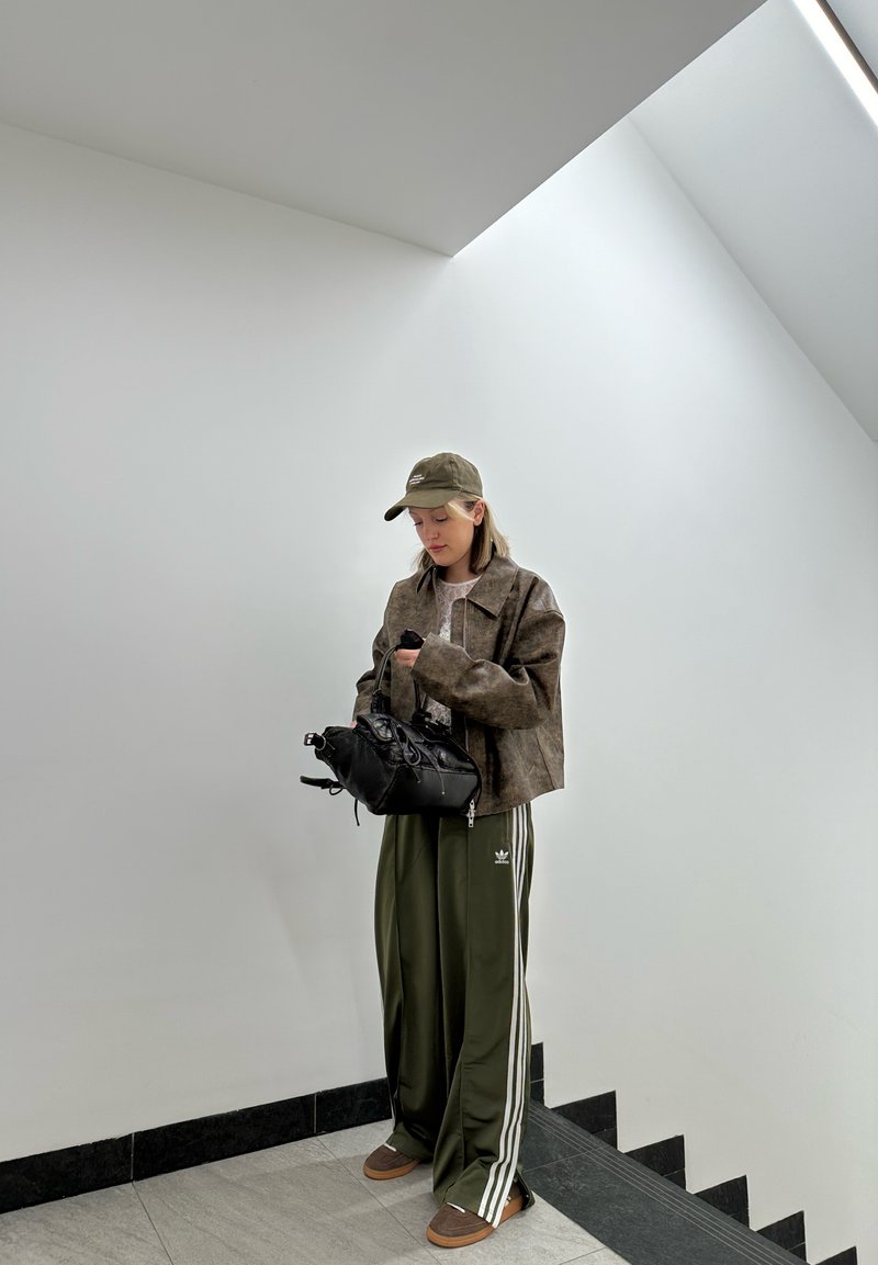 Καφέ κοντό σακάκι, ελαιόχρωμο παντελόνι με άσπρες ρίγες, μαύρη τσάντα και σκούρα sneakers. Ο χώρος χαρακτηρίζεται από έναν μινιμαλιστικό λευκό τοίχο.