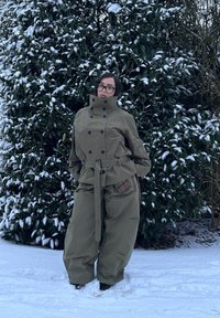 Un vêtement d'extérieur vert surdimensionné avec un col montant, un design croisé à double boutonnage et une poche marron à carreaux ; porté avec un pantalon ample.