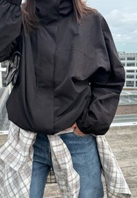 Veste noire, légère, avec un col haut et des poignets élastiques, associée à un jean bleu et une chemise à carreaux nouée autour de la taille.