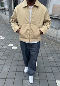 Beige zip-up jack met twee voorzakken; witte textuurshirt eronder; donkere spijkerbroek; witte sneakers; staand op een betegeld trottoir.