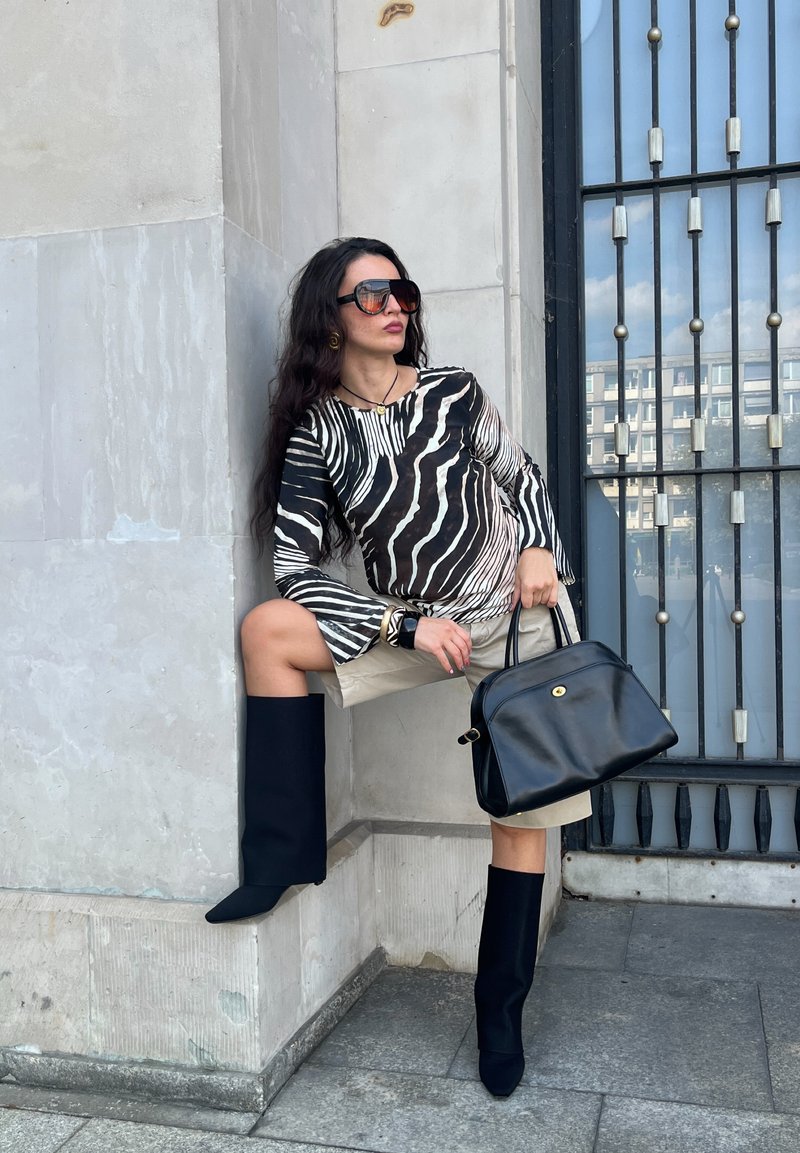 Blouse à motifs zèbre noir et blanc avec des manches évasées, short beige, bottes noires montantes et un sac à main en cuir noir avec un accent doré.