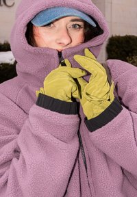 Personne portant une casquette bleue, une veste polaire violette fermée jusqu'au menton, ajustant des gants jaunes vifs en plein air.