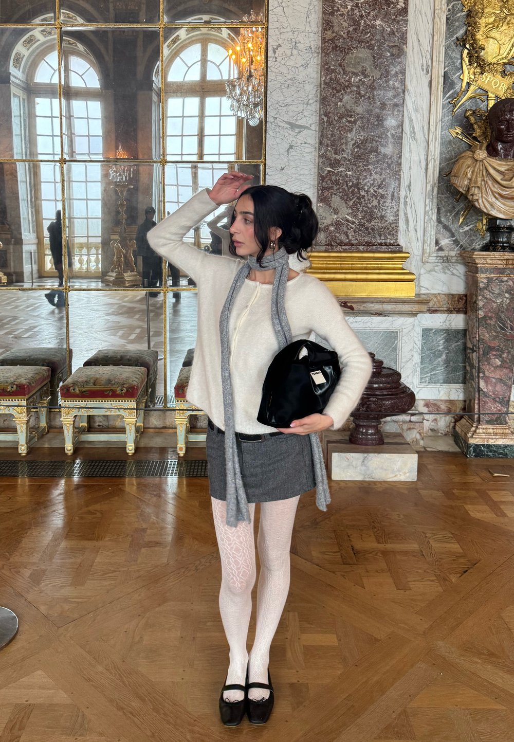 Femme portant un pull blanc, une jupe grise et des collants à motifs, posant avec un sac noir à l'intérieur d'une pièce ornée de colonnes en marbre et de grands miroirs.