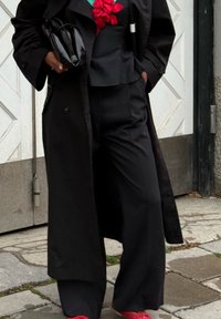 Άτομο που φοράει μαύρο παντελόνι με φαρδιά ποδόγυρα, μαύρο παλτό, μαύρο τοπ με κόκκινη λεπτομέρεια λουλουδιού, κρατώντας μαύρο τσαντάκι, περπατώντας πάνω σε λιθόστρωτο δρόμο.