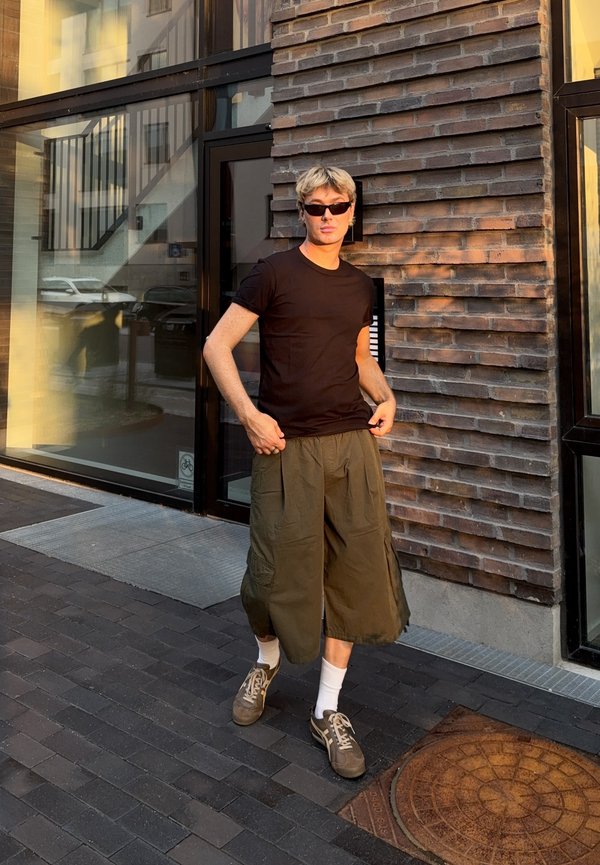 Jong persoon met kort blond haar draagt een zonnebril, een zwart t-shirt, brede olijfgroene broek en sneakers, staat bij een bakstenen muur in het zonlicht.