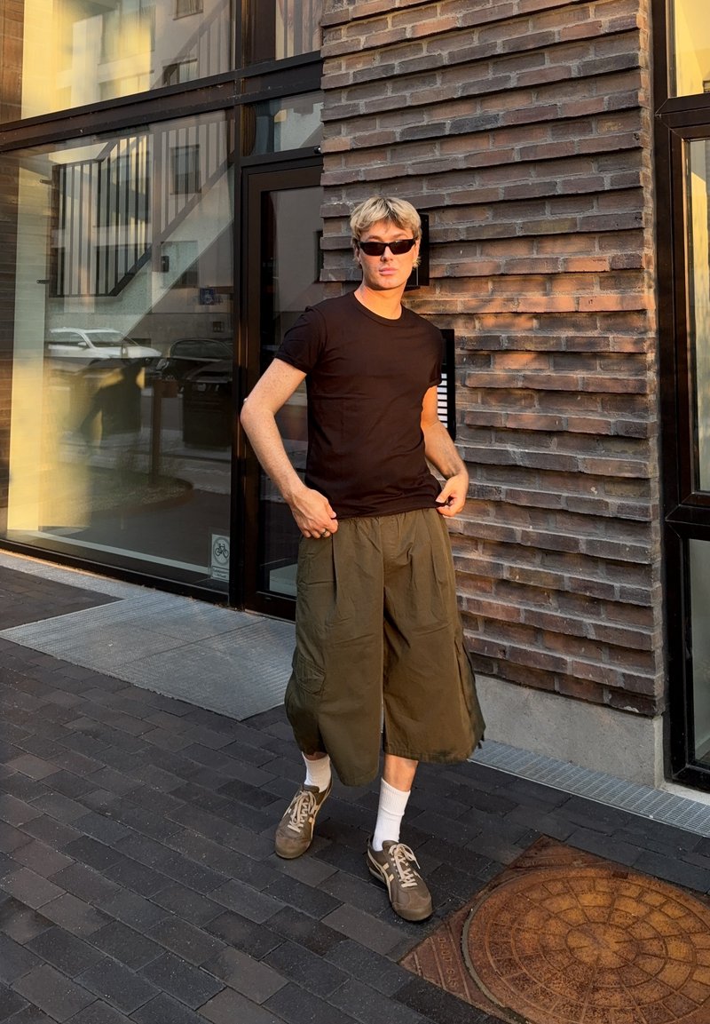 Jeune personne aux cheveux blonds courts portant des lunettes de soleil, un t-shirt noir, un pantalon large kaki et des baskets, debout près d'un mur en briques au soleil.