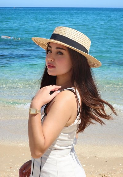 Sombrero de paja de ala ancha con una banda negra, vestido sin mangas de color claro con tirantes finos, reloj dorado, con un fondo de playa y océano.