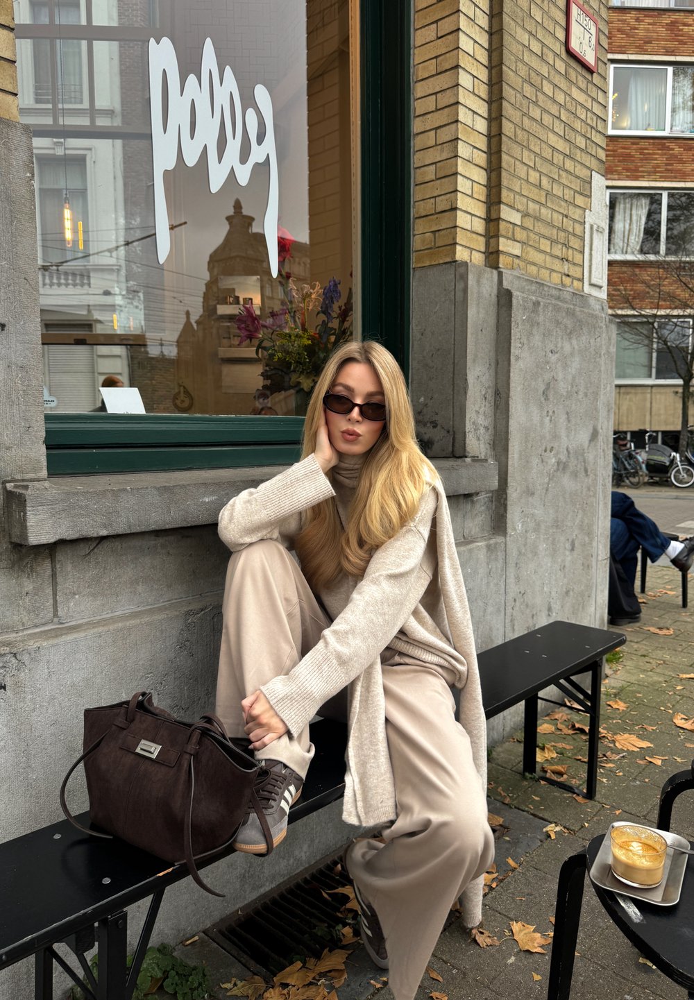 Uma pessoa sentada com um suéter e calças bege, usando óculos escuros, com uma bolsa castanha e ténis cinzentos, ao lado de um café numa bandeja.