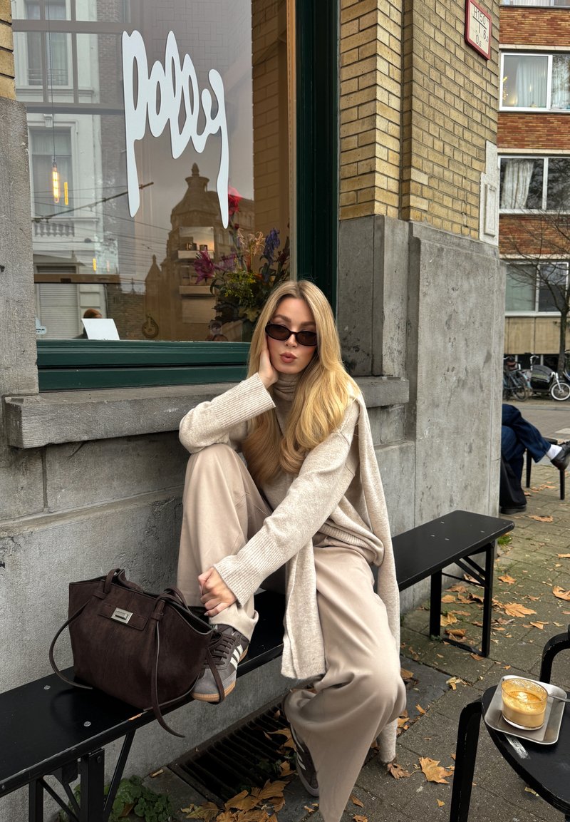 A seated person in a beige sweater and pants, wearing dark sunglasses, with a brown bag and gray sneakers, beside a coffee on a tray.