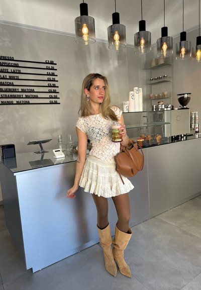 Blusa de encaje, falda plisada crema con borde de encaje, medias marrones, botas beige tipo slouchy, sosteniendo una bebida, con un fondo de café minimalista.