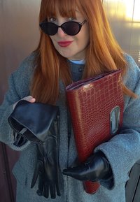 Kobieta z czerwonymi włosami i czerwoną pomadką, nosząca czarne okulary przeciwsłoneczne i szary płaszcz, trzyma czarne skórzane rękawiczki oraz czerwony fakturowany kopertówkę.