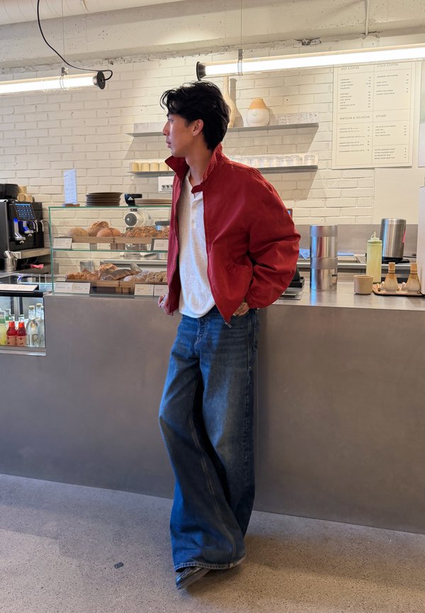 Mann in roter Jacke und weitem Jeans steht mit den Händen in den Taschen an einem Caféthekenbereich, der Gebäck und Kaffeemenü auf einer weißen Backsteinwand zeigt.