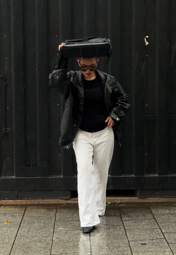 Une personne portant des lunettes de soleil, une veste en cuir noire et un pantalon blanc passe devant une entrée sombre, en tenant un sac noir plat au-dessus de sa tête.