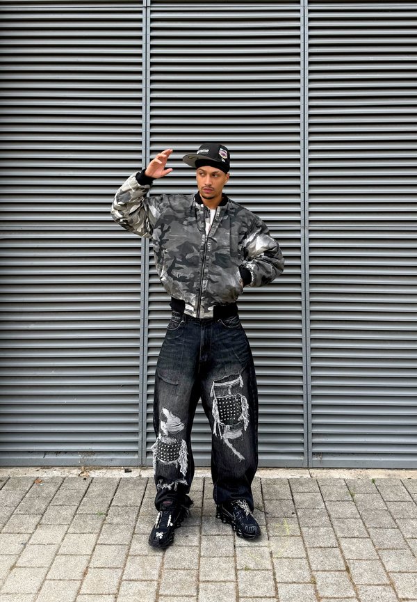 Homme portant une veste camouflage, une casquette noire et un jean noir déchiré, debout sur une surface pavée devant un mur métallique de ventilation.