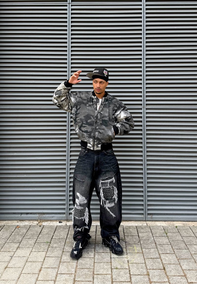 Homme portant une veste camouflage, une casquette noire et un jean noir déchiré, debout sur une surface pavée devant un mur métallique de ventilation.