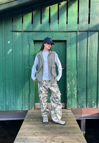 Pantalones cargo de patrón camuflaje, camisa de manga larga gris claro, chaleco verde y gorra oscura. De pie sobre una plataforma de madera frente a un fondo verde.