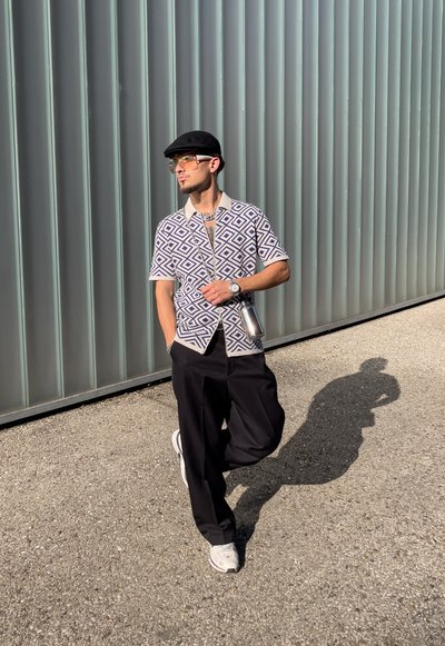 Camisa de manga corta con un patrón geométrico en azul marino y crema, combinada con pantalones negros y zapatillas blancas. Fondo de paneles metálicos verticales.