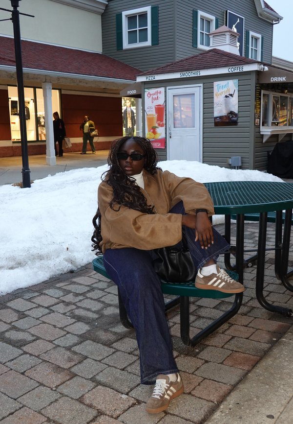 Henkilö, jolla on aurinkolasit, ruskea takki, siniset farkut ja tennarit, istuu ulkona vihreällä metallipöydän penkillä lumisen alueen ja ruokakojun lähellä.