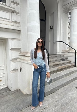 Mujer con gafas de sol, top azul claro transparente, vaqueros acampanados y tacones puntiagudos, sosteniendo un bolso plateado, parada junto a escalones y columnas de piedra.