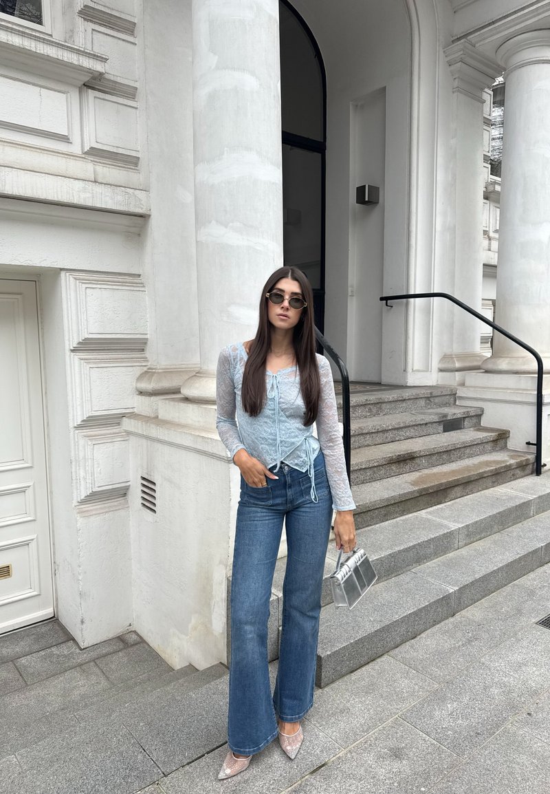 Γυναίκα με γυαλιά ηλίου, διάφανο ανοιχτόμπλε τοπ, καμπάνα τζιν και μυτερές γόβες, κρατώντας μια ασημένια τσάντα, στέκεται δίπλα σε πέτρινα σκαλοπάτια και κολόνες.