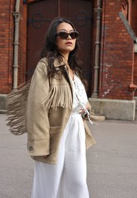 Bežs frizēts jaka ar maigu tekstūru, apvienota ar baltu blūzi un platām biksēm. Saulesbrilles ir kaķa acs formas ar tumšu rāmīti.