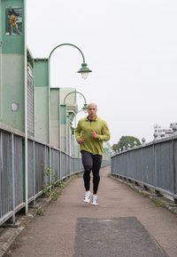 Man i grön jacka och svarta leggings joggar på en inhägnad väg under böjda gatlyktor en molnig dag.