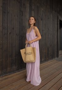 Ανοιχτό ροζ, αμάνικο μάξι φόρεμα με χαλαρή γραμμή. Κρατάει μια μπεζ τσάντα με μαύρο κείμενο. Ξύλινο φόντο και δάπεδο από σανίδες.