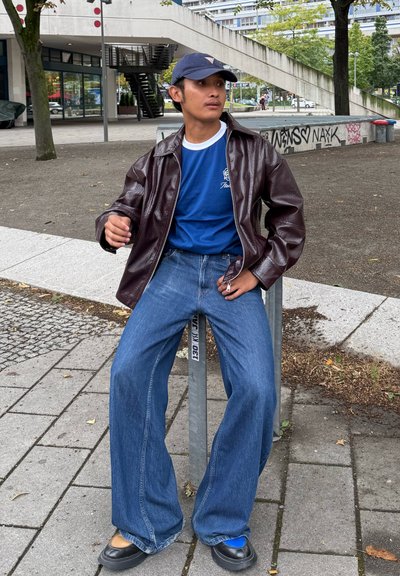 Chaqueta de charol marrón sobre una camiseta azul, jeans de pierna ancha azules y zapatos negros. Sentado en un soporte para bicicletas en un entorno urbano.
