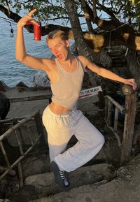 Pelēka bez piedurknēm krekls un baltas vaļīgas bikses, ko valkā persona ar sarkanu bundžu rokā, pozējot pie kāpnēm pludmales apkārtnē.