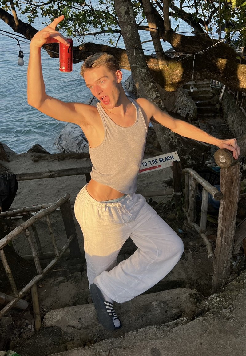 Grijs mouwloos overhemd en witte losse broek gedragen door een persoon die een rode blik vasthoudt, poserend bij een trap in een strandomgeving.