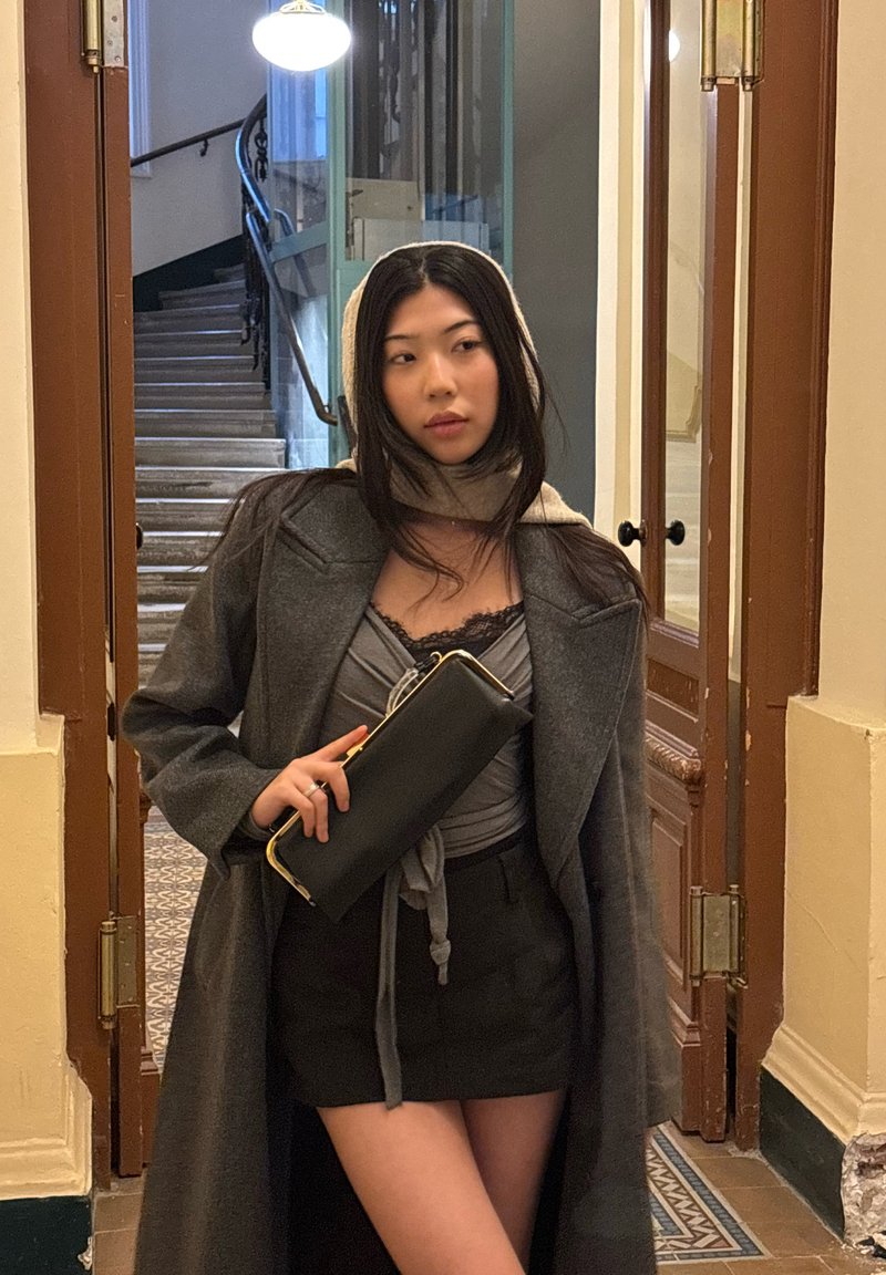 Long manteau gris, pochette noire, haut gris avec détail à nouer. Debout dans un couloir avec des portes en bois et des escaliers visibles en arrière-plan.