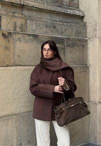 Rjav volnen plašč z visokim ovratnikom in širokimi rokavi, kombiniran s temno rjavo semišno torbico z zlatimi kovinskimi dodatki. Videzu dodajo bel hlače.