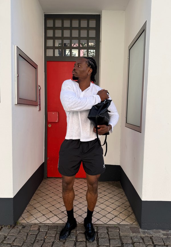 Hombre con camisa blanca de manga larga y pantalones cortos negros, de pie en una puerta estrecha con una puerta roja, sosteniendo una bolsa negra.