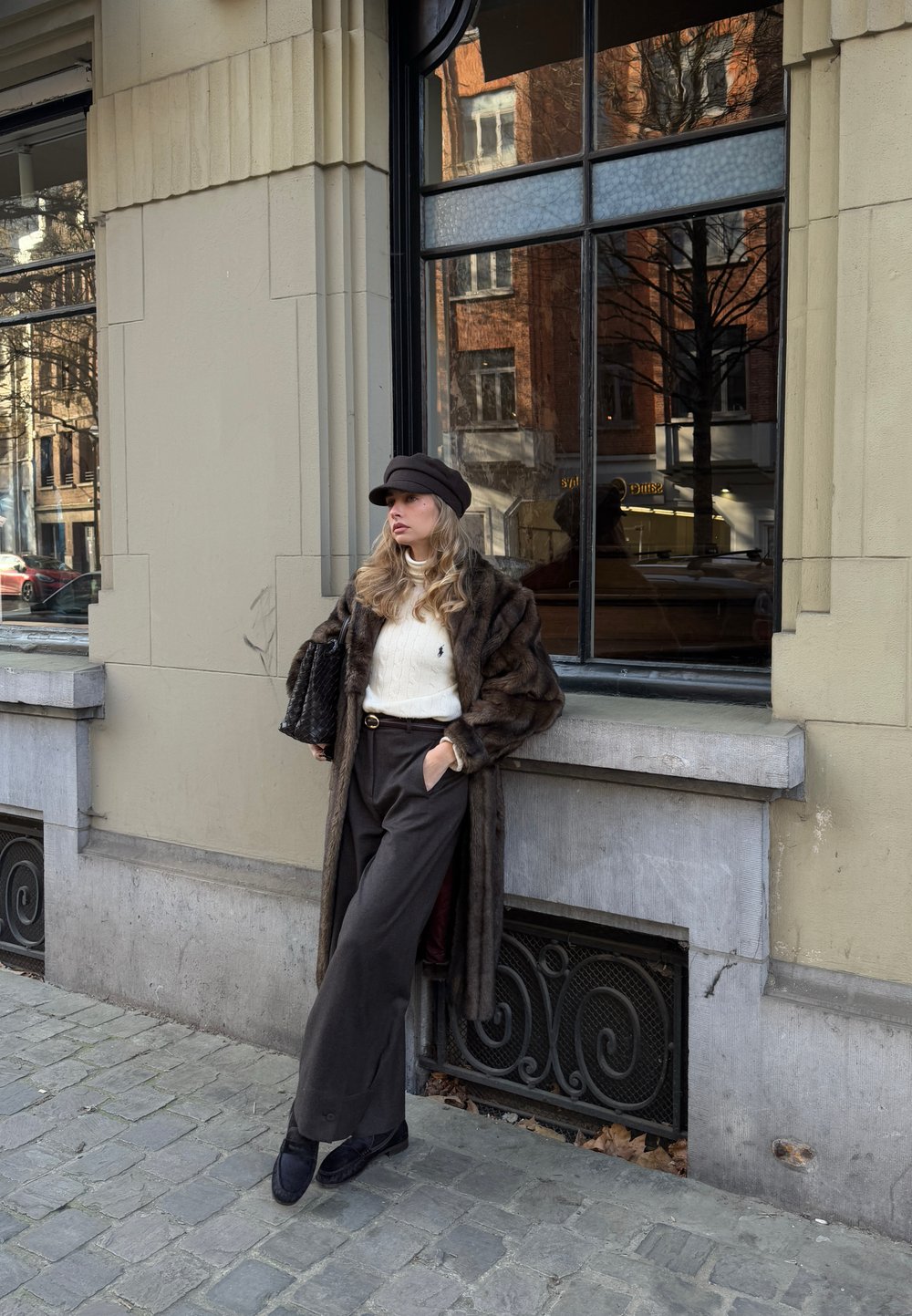 Νεαρή γυναίκα με καφέ γούνινο παλτό, λευκό πουλόβερ και μαύρο καπέλο, στηριζόμενη σε πέτρινο τοίχο σε πλακόστρωτο δρόμο κοντά σε μεγάλο παράθυρο.