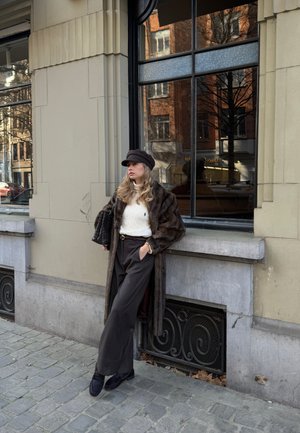 Joven mujer con abrigo de piel marrón, suéter blanco y gorra negra apoyada contra una pared de piedra en una calle empedrada cerca de una ventana grande.