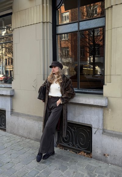 Joven mujer con abrigo de piel marrón, suéter blanco y gorra negra apoyada contra una pared de piedra en una calle empedrada cerca de una ventana grande.